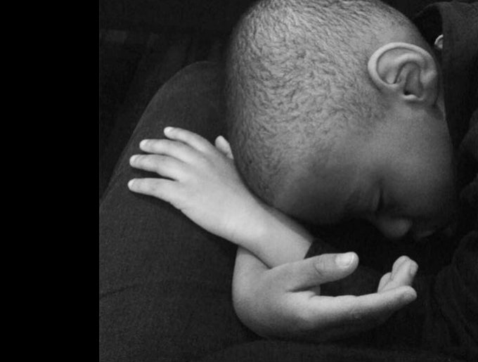 Black and white photograph of a young Black boy resting his head in his crossed hands on someone’s lap. His eyes are closed