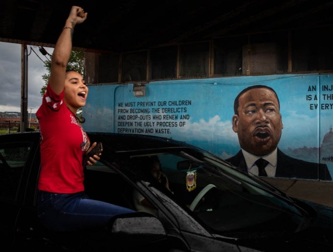 Horizontal format photograph of a person with a raised fist leaning out of a car window. A mural depicting Martin Luther King, Jr. is in the background